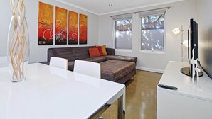 A modern living room with a white table, a brown sofa, and a TV on a stand. There are abstract paintings on the wall and a window with blinds. - Hurtle Sq Hideaway - Parking, Central, Ground Flr (Adelaide)