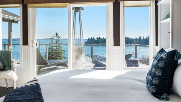 A bedroom with a large bed, a view of the water, and a balcony with chairs. - Epic Views: Hot Tub, 3BR/3BA, Sleeps10, Remote Retreat! (Lakebay)