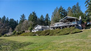 A two-story house with a large deck and a covered porch, surrounded by a well-maintained lawn and tall trees. - Epic Views: Hot Tub, 3BR/3BA, Sleeps10, Remote Retreat! (Lakebay)