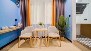 Una habitación pequeña y moderna con una pared azul, una mesa de madera con dos sillas, una planta pequeña y una cocina visible a través de una puerta abierta.