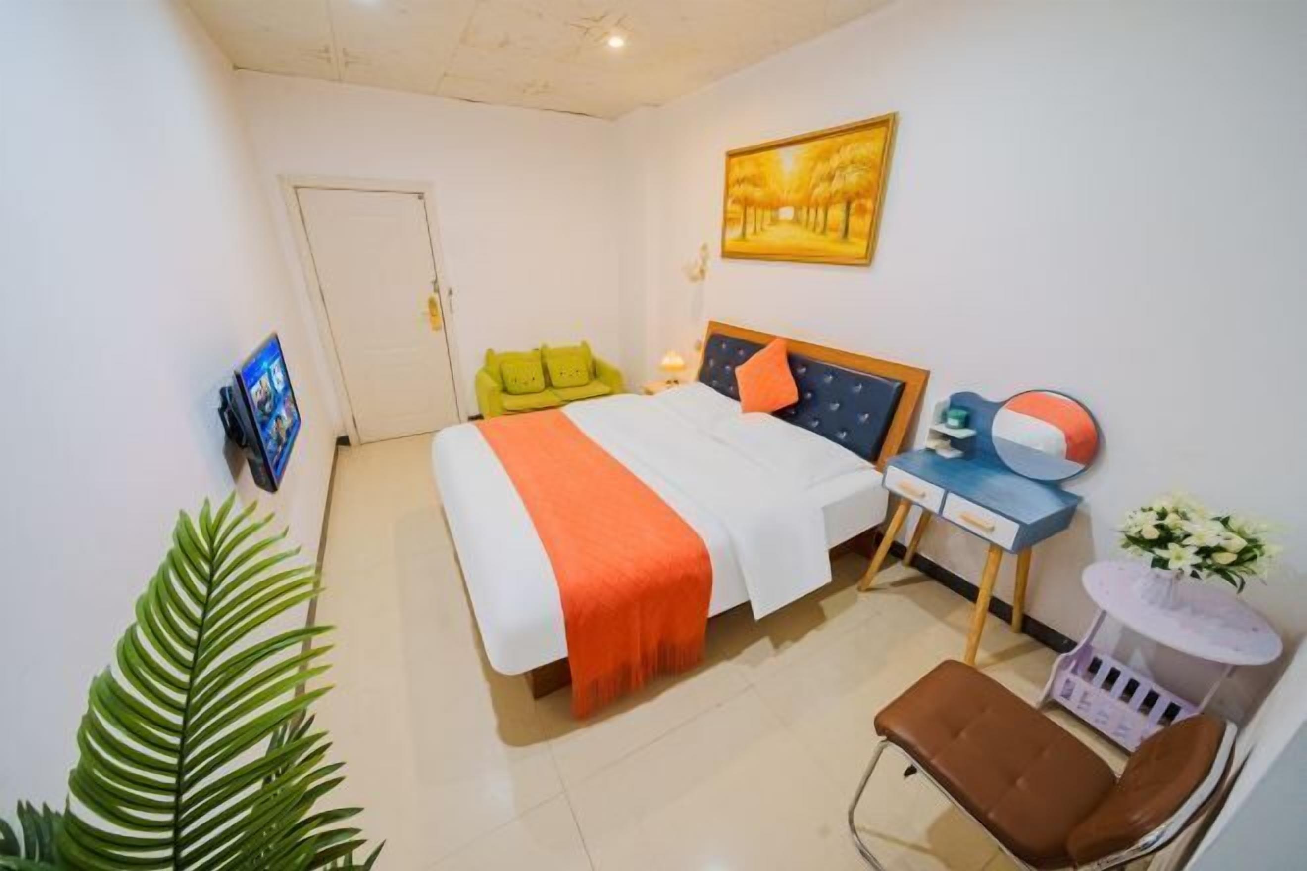 A hotel room with a bed, a desk with a chair, a small table with flowers, and a large plant.