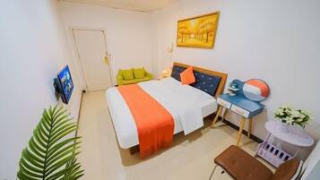 A hotel room with a bed, a desk with a chair, a small table with flowers, and a large plant.