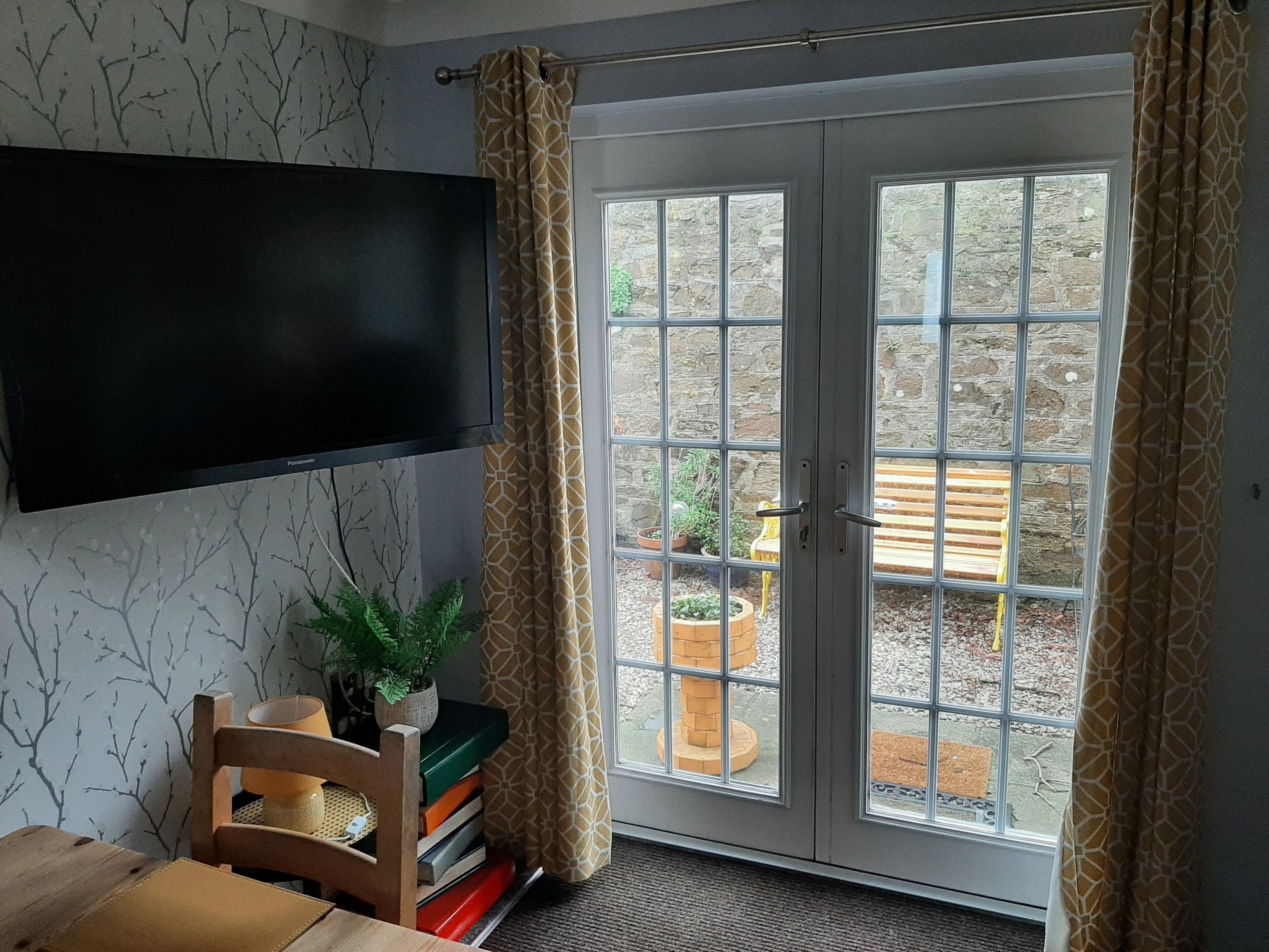 A room with a television mounted on the wall, a chair, a small table, and a bookshelf. There is a sliding glass door leading to an outdoor area with a bench and potted plants.