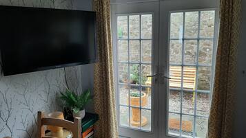A room with a television mounted on the wall, a chair, a small table, and a bookshelf. There is a sliding glass door leading to an outdoor area with a bench and potted plants.