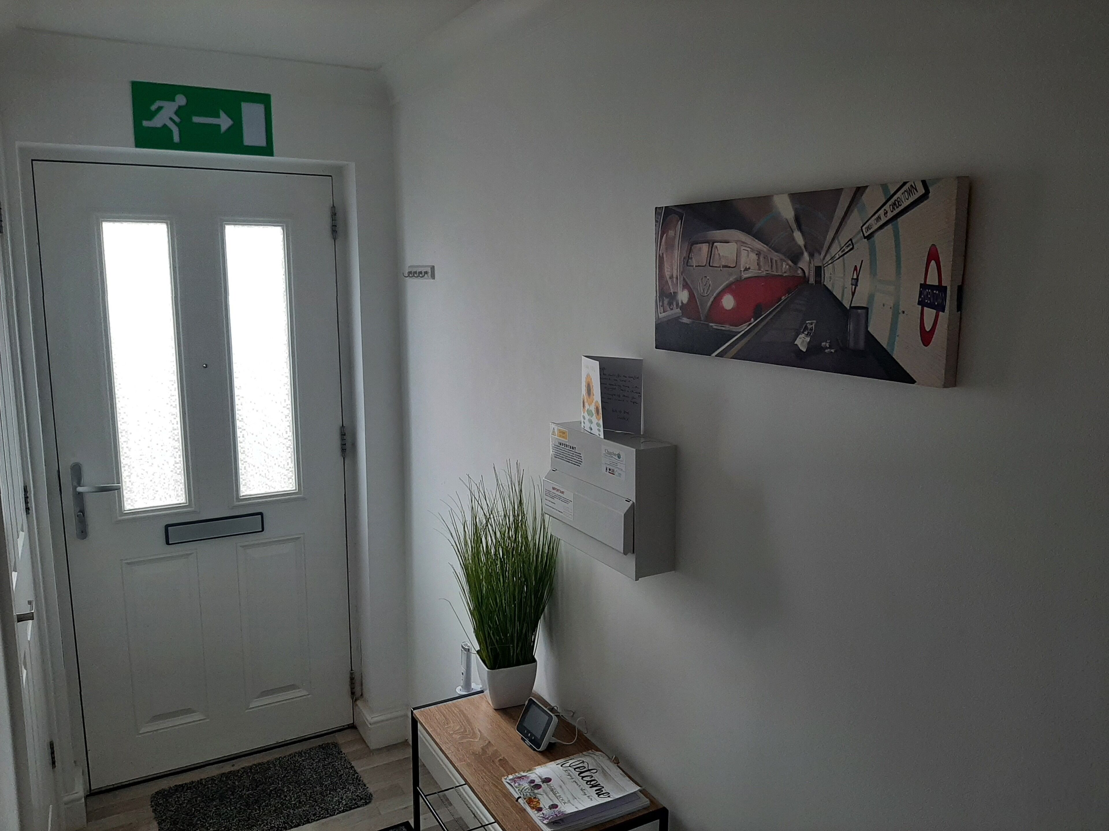 Un espace d’accueil avec une porte blanche, un panneau vert indiquant la sortie, un paillasson, une plante en pot, un panneau d’information fixé au mur et une œuvre d’art encadrée.