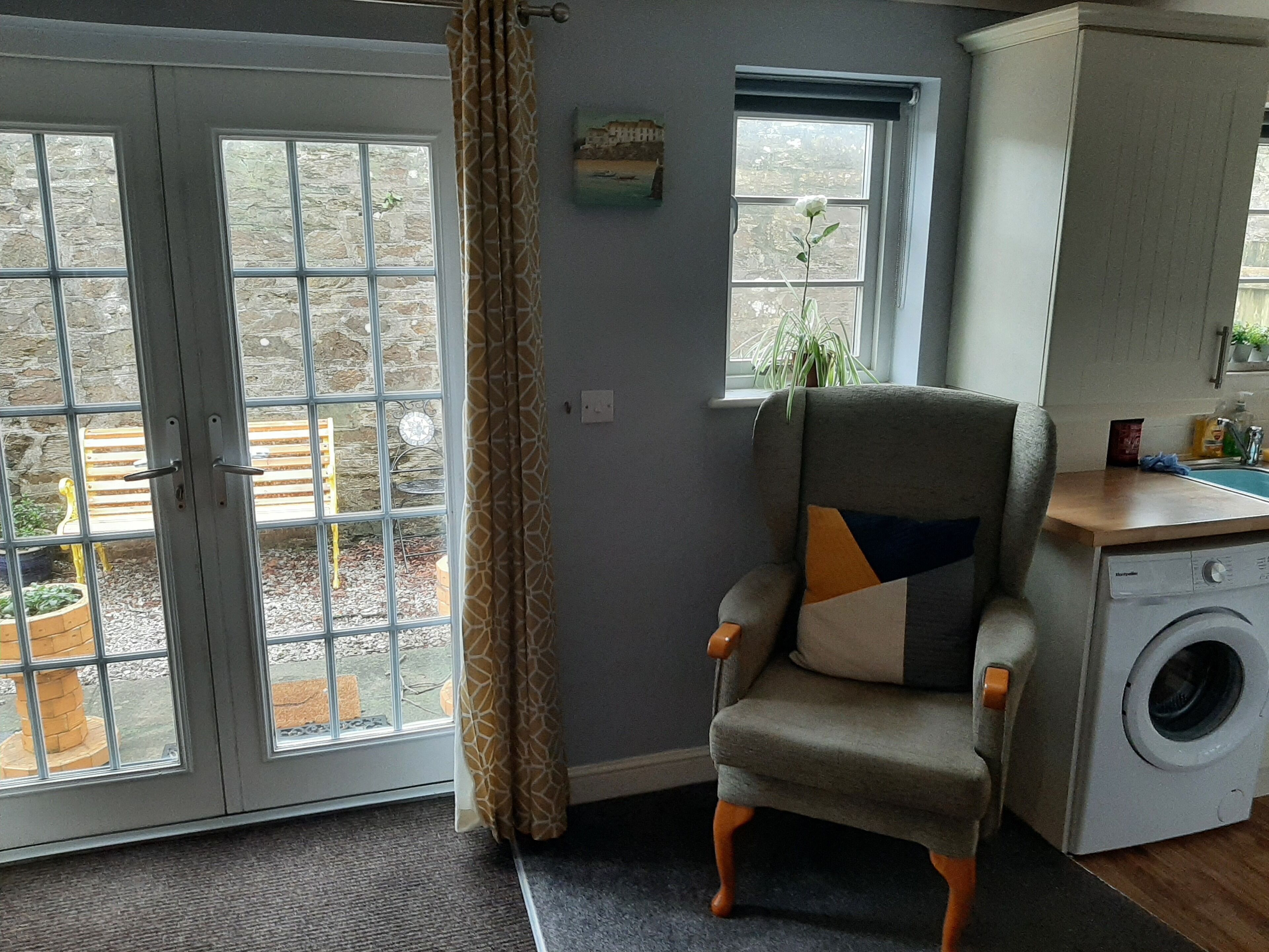 A room with a grey armchair, a washing machine, and a view of an outdoor area with a bench and a potted plant.