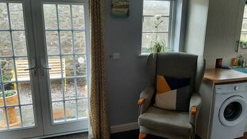 A room with a grey armchair, a washing machine, and a view of an outdoor area with a bench and a potted plant.