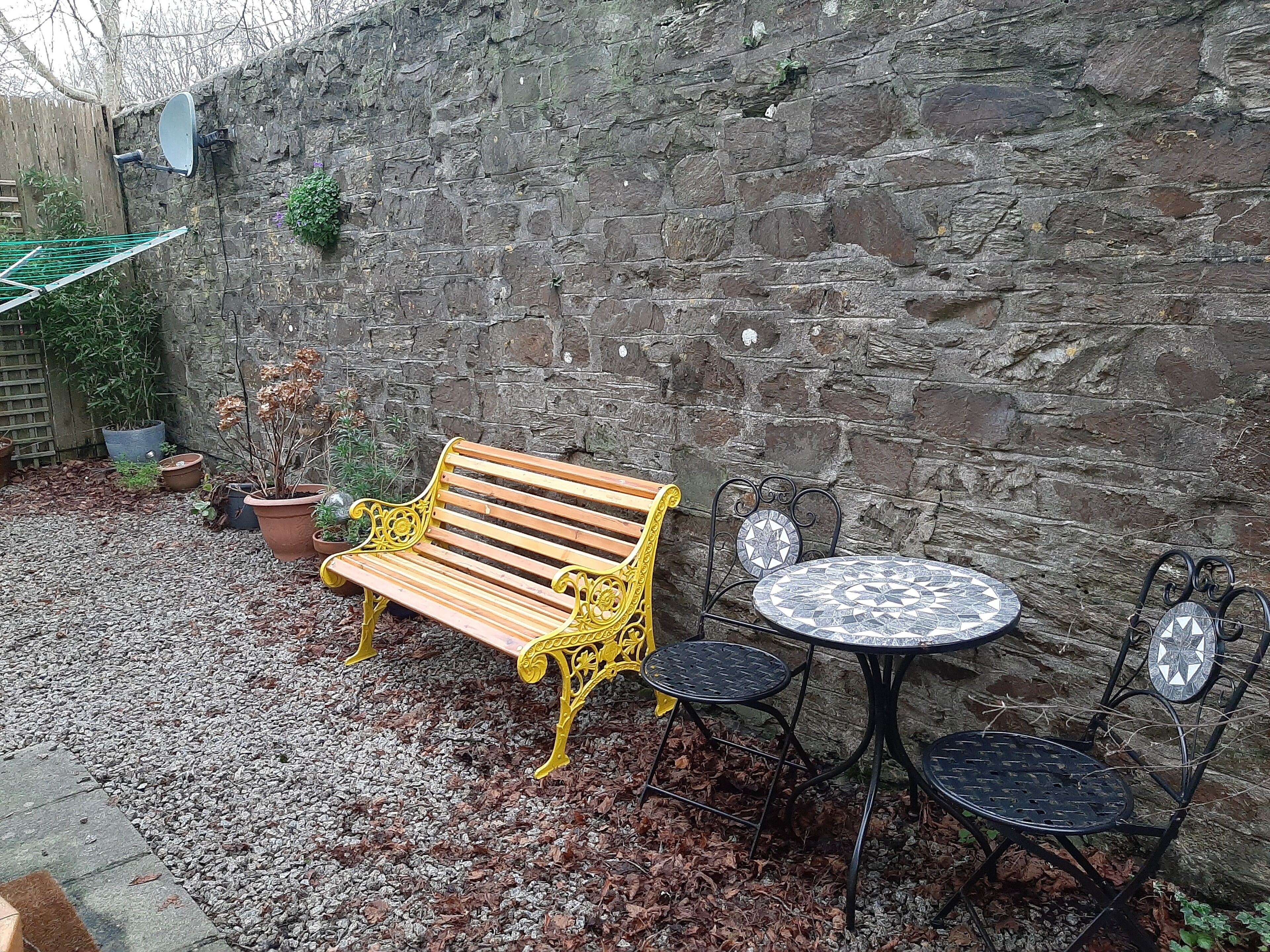 A wooden bench with yellow metal legs and a metal table with chairs in a courtyard with a stone wall.