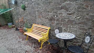 A wooden bench with yellow metal legs and a metal table with chairs in a courtyard with a stone wall.