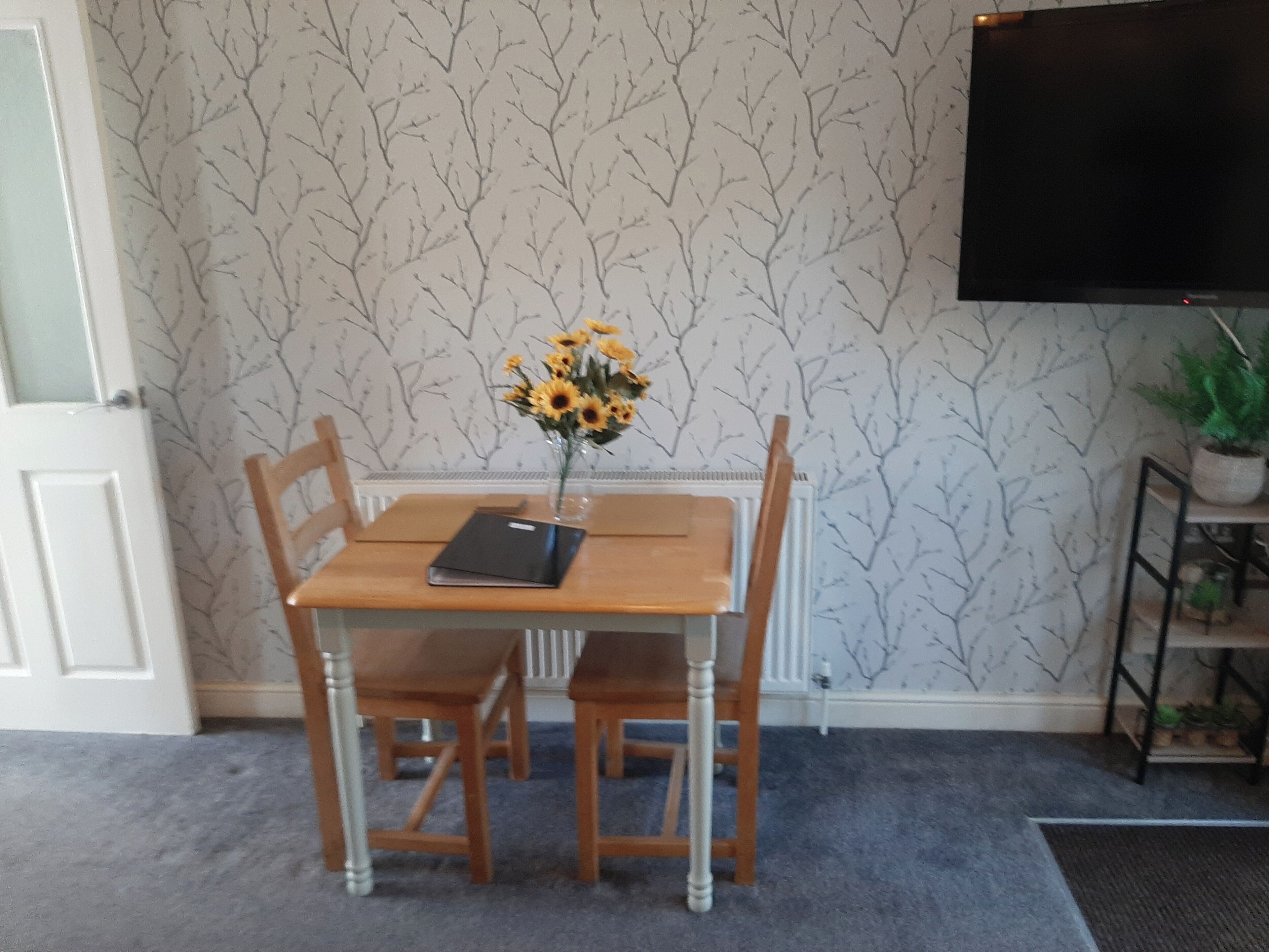 A dining area with a table, chairs, a vase with sunflowers, a black folder, and a TV on the wall.