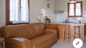 A living room with a brown leather sofa, a wooden bar counter with stools, and a kitchen area with white cabinets and a stove. - Villa Delphia One Bedroom Traditional House (Kefalonia)