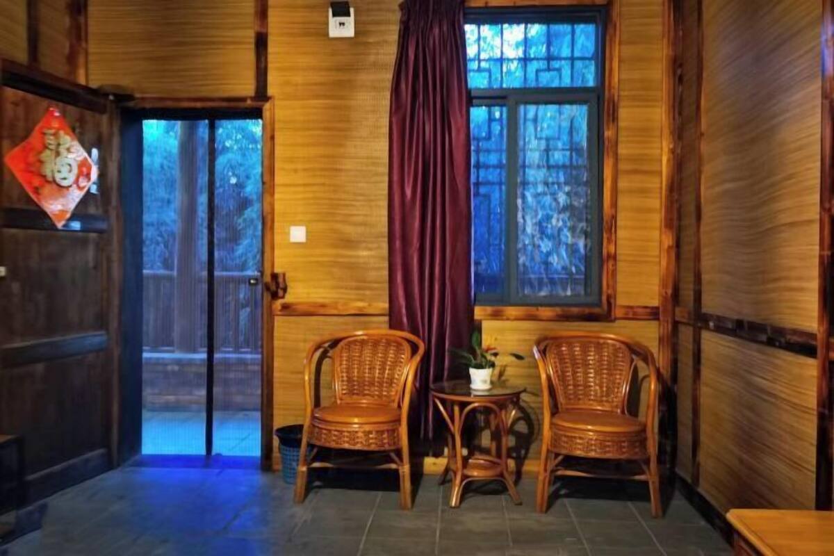 Une pièce aux murs en bois, une grande fenêtre donnant sur des arbres, deux fauteuils en osier, une petite table avec une plante et un rideau rouge.