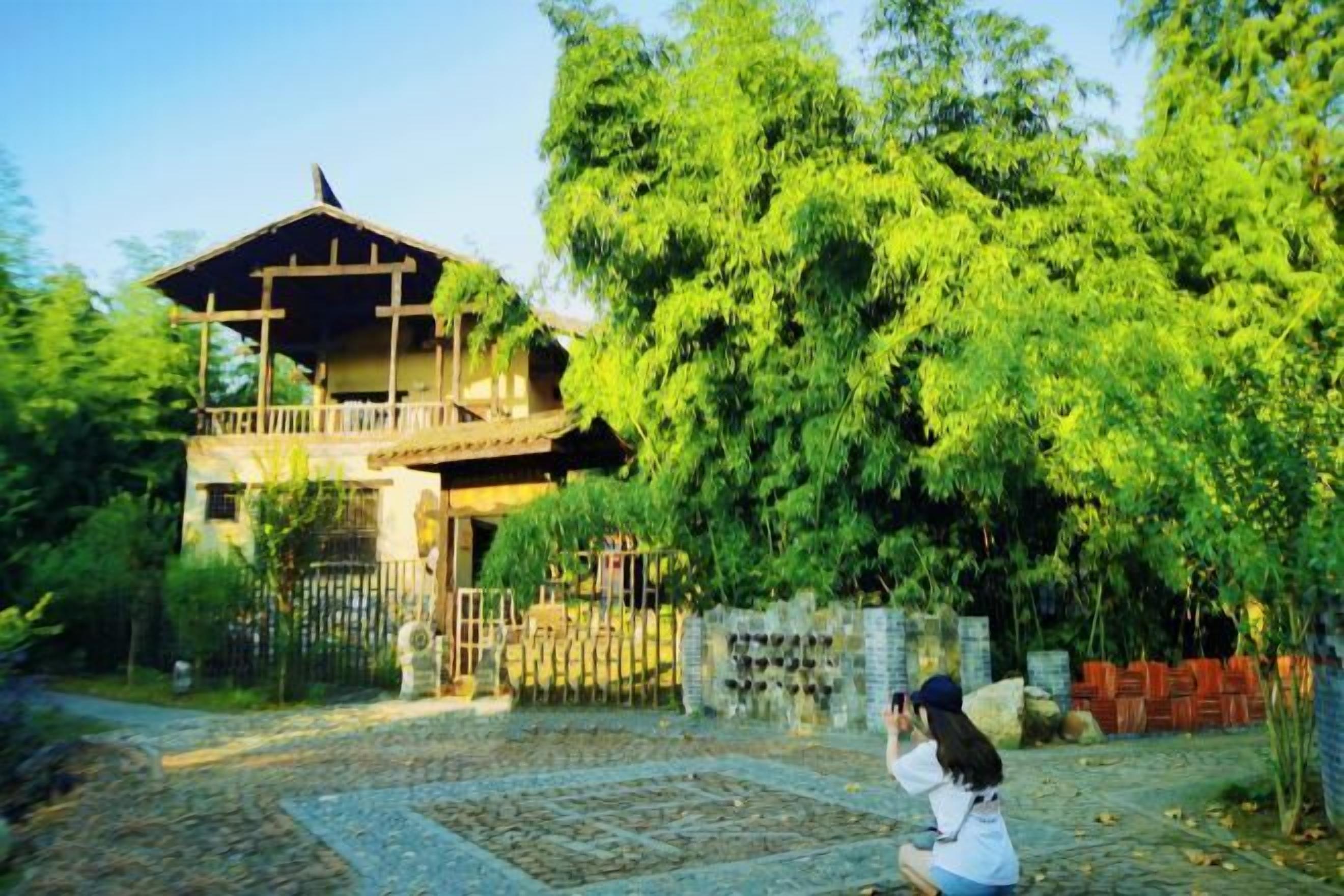 A traditional wooden house with a balcony, surrounded by lush greenery and a stone pathway.