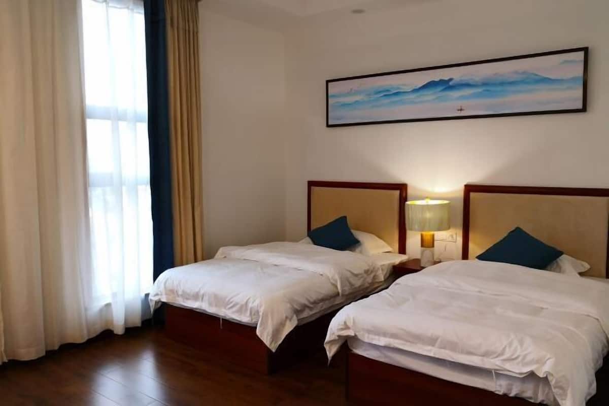 Une chambre d’hôtel avec deux lits, une grande fenêtre avec des rideaux, un tableau au mur et un plancher en bois.