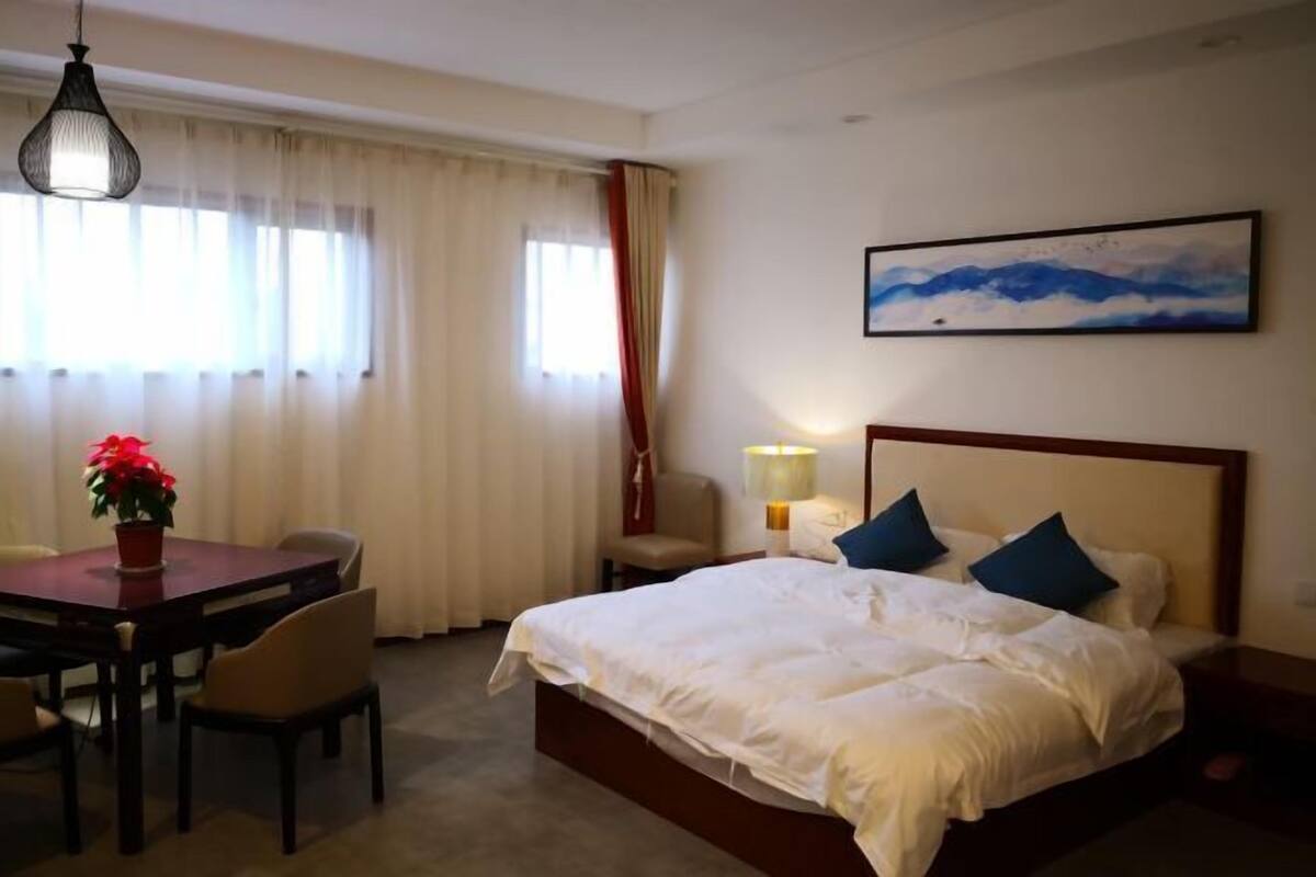 Une chambre d’hôtel avec un lit, un bureau avec des chaises, une table avec un pot de fleurs, un tableau au mur et une fenêtre avec des rideaux.