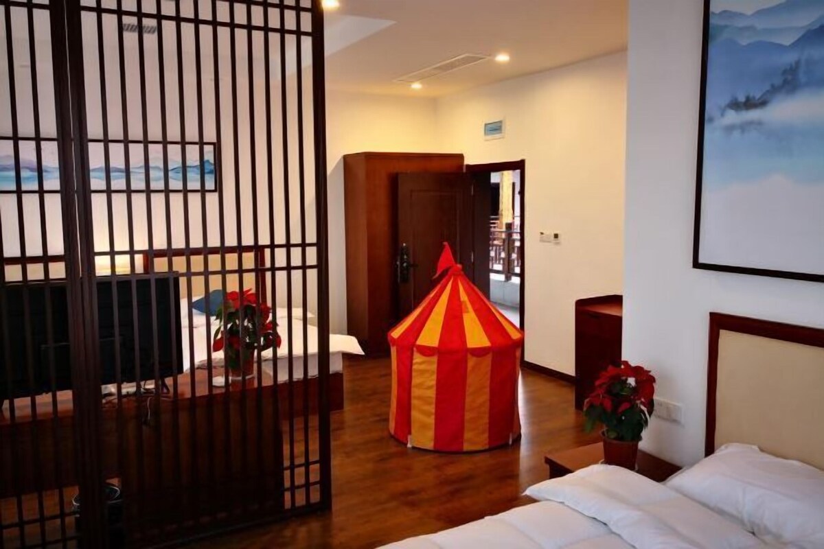 A hotel room with a bed, a desk, a chair, a small red and yellow tent, and a painting on the wall.
