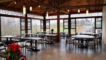 A spacious indoor area with tables and chairs arranged for dining, large windows with a view of the outdoors, and a wooden ceiling structure.