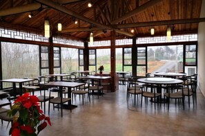 A spacious indoor area with tables and chairs arranged for dining, large windows with a view of the outdoors, and a wooden ceiling structure. - Huaqitang Rural Life Hostel (Wuhan)