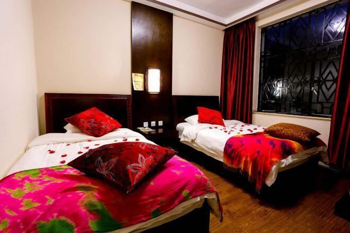 Une chambre d’hôtel avec deux lits simples, chacun agrémenté de draps rouges et roses, d’une tête de lit en bois et d’une fenêtre ornée d’une grille métallique.