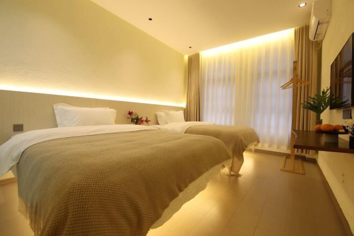 A hotel room with two beds, a bedside table with a lamp, a large window with curtains, and a wall-mounted air conditioner.