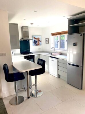 A compact kitchen with a dining area, white cabinets, a stainless steel refrigerator, and a window with blinds. - The Cart House - Staverton Cheltenham (Cheltenham)
