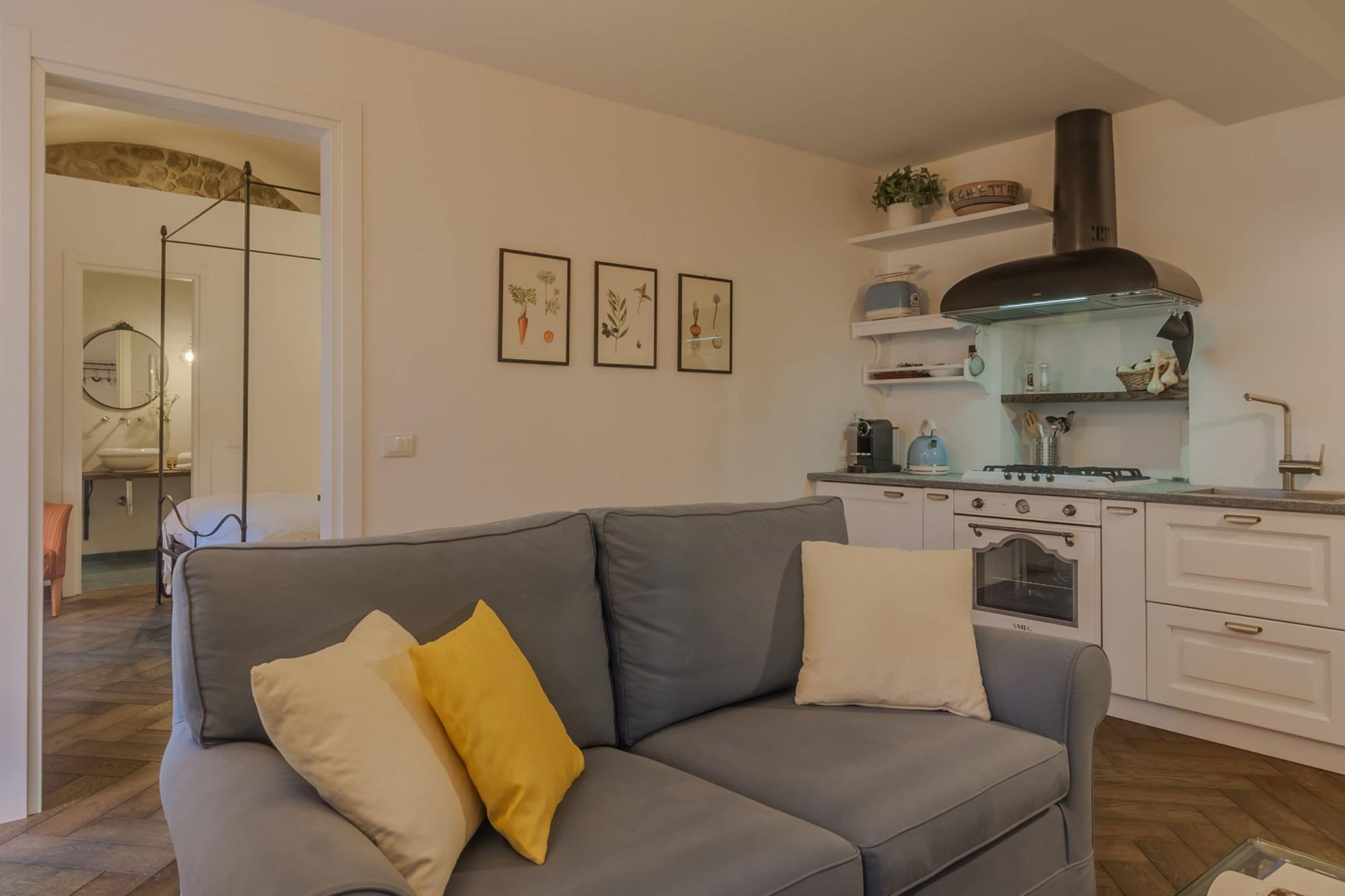 A modern kitchen with white cabinets, a stove, and a black range hood. There is a gray sofa with yellow and white cushions in the foreground.