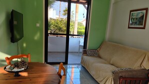 A room with a wooden dining table, a sofa, and a television mounted on the wall. - Lovely 1-bed Palm Garden House in Perivolialarnaka (Pervolia)