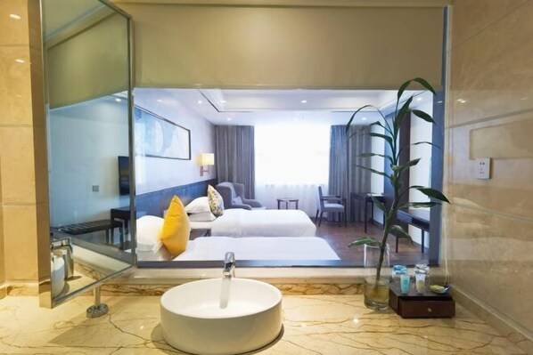 Ein Hotelbad mit einem großen Spiegel, einem runden Waschbecken und einem Bett, das durch die Glasabtrennung sichtbar ist.