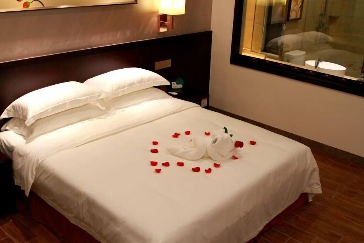 Quarto de hotel com cama bem arrumada, mesa de cabeceira com abajur e janela com vista para o banheiro.