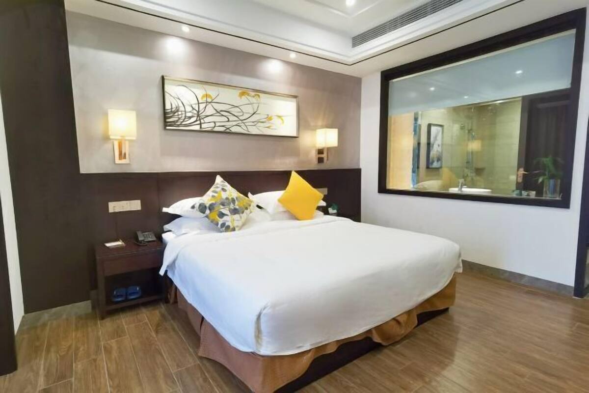 Quarto de hotel com uma cama grande, uma mesinha de cabeceira, uma obra de arte na parede e uma janela ampla.