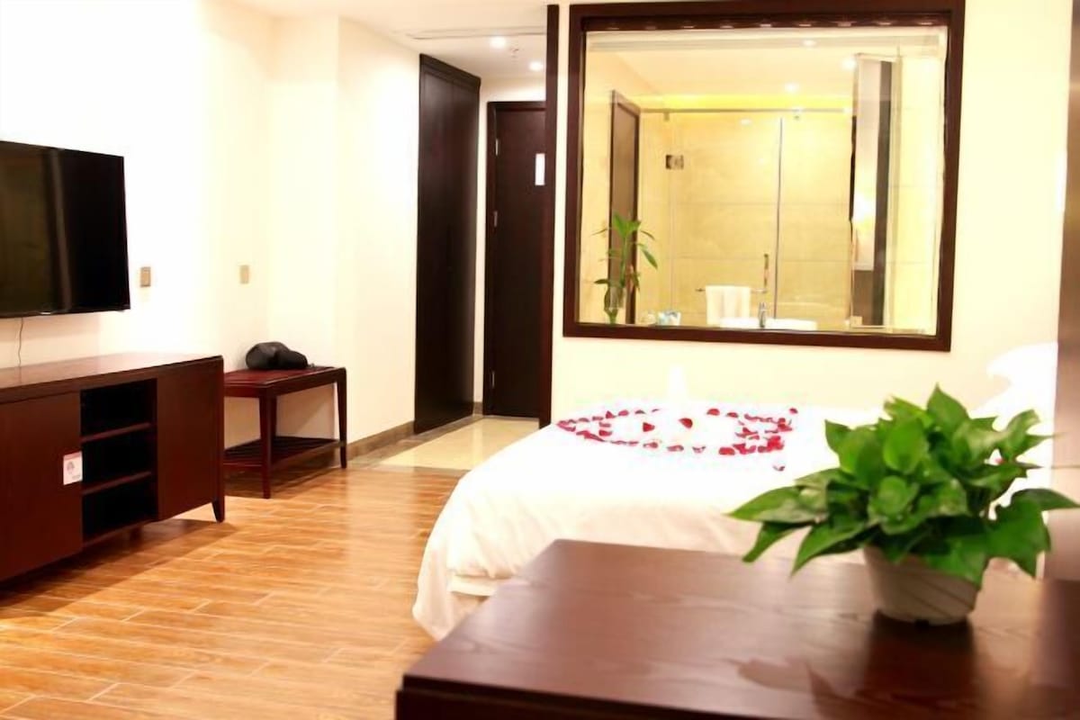 A hotel room with a bed, a television, a small table, a potted plant, and a large mirror reflecting a bathroom.