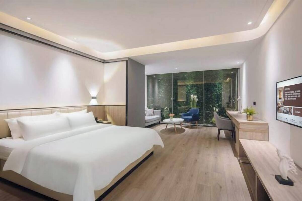Ein modernes Hotelzimmer mit einem großen Bett, einem Schreibtisch und einer Sitzecke mit Blick auf Grünanlagen durch eine Glaswand.