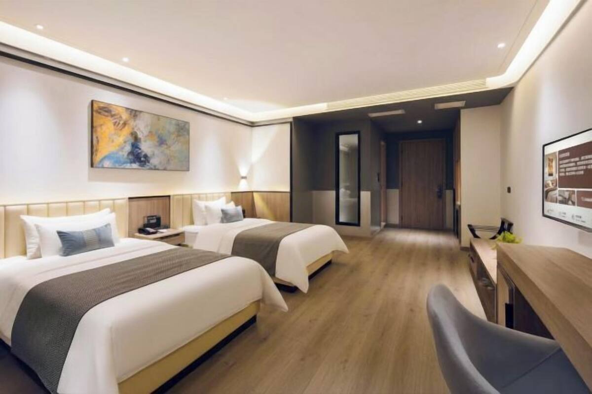 Ein Hotelzimmer mit zwei Betten, einem Schreibtisch und einem Stuhl. An der Wand hängt ein Bild, und eine Tür führt in einen anderen Raum.