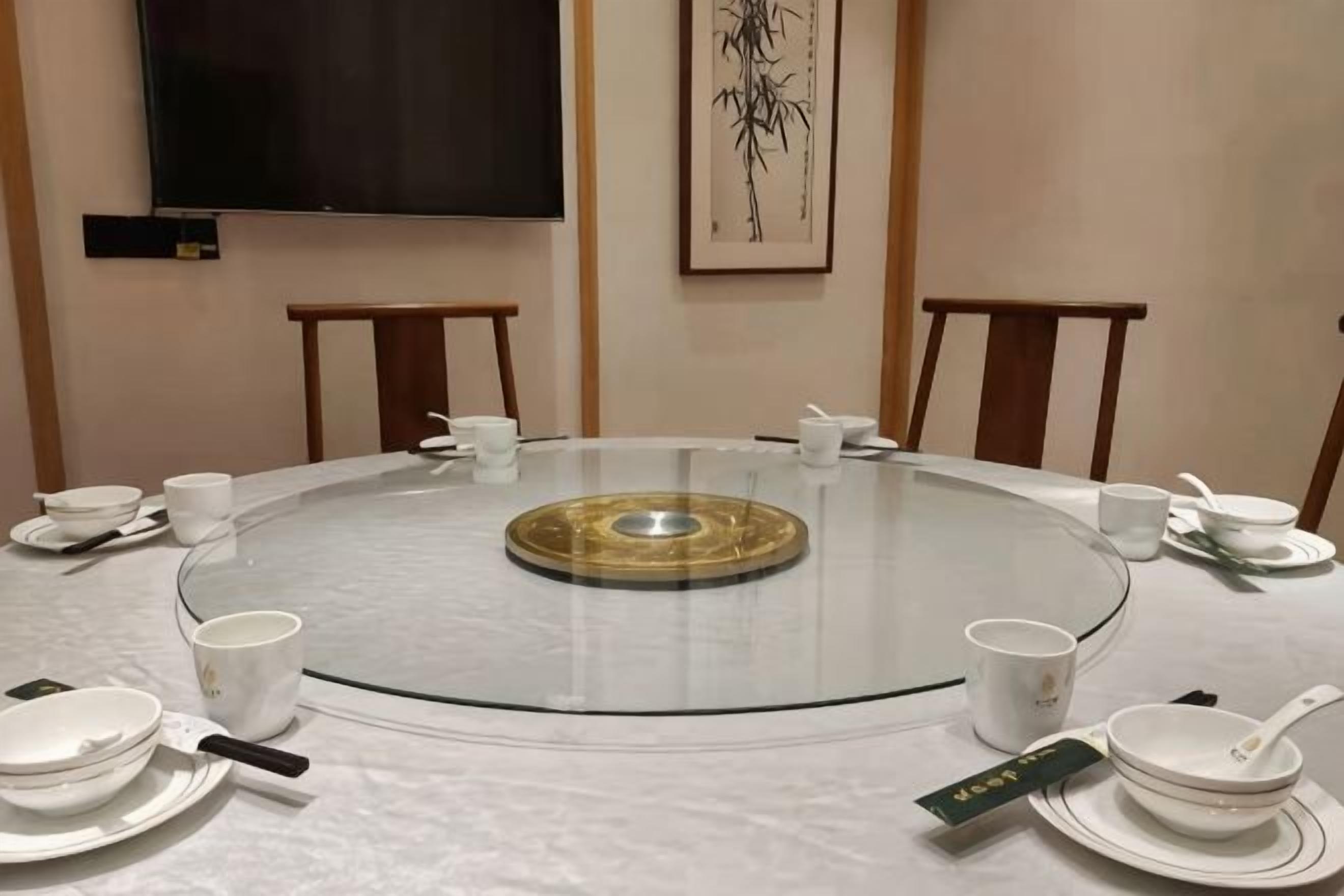 A round glass table with white plates, cups, and a tea set, surrounded by chairs in a room with a flat-screen TV and a framed artwork on the wall.