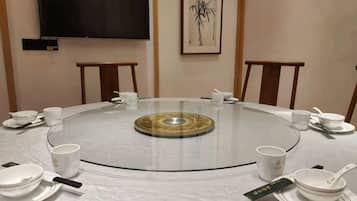 A round glass table with white plates, cups, and a tea set, surrounded by chairs in a room with a flat-screen TV and a framed artwork on the wall.