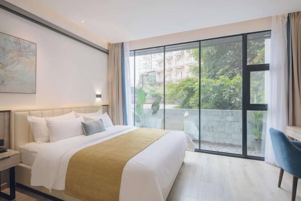 Ein Hotelzimmer mit einem großen Bett, einem Schreibtisch, einem Stuhl und einem Balkon mit Blick auf Gebäude und Grünflächen.