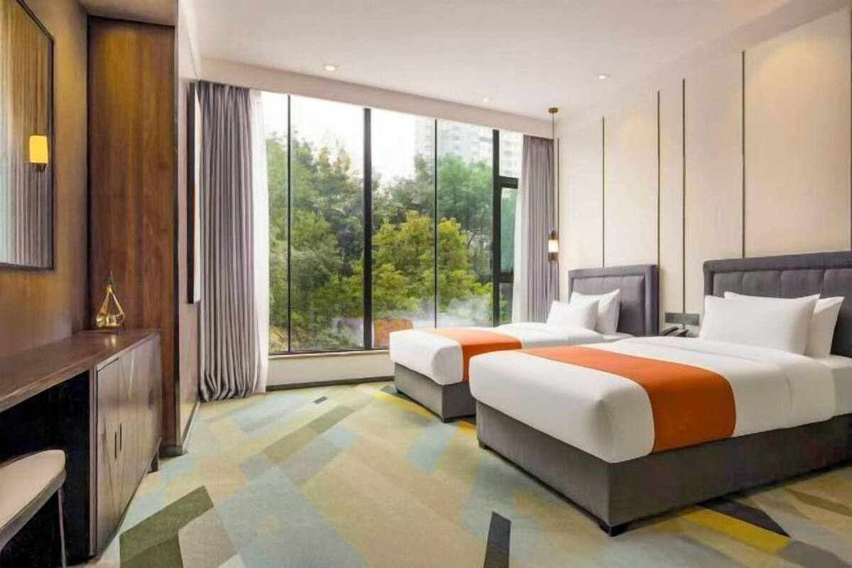 Ein Hotelzimmer mit zwei Betten, einem Schreibtisch und einem großen Fenster mit Blick auf Bäume.