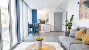 Un salon moderne avec un canapé, une table basse ornée d’un vase de fleurs, et une vue sur la cuisine et la salle à manger grâce à de grandes fenêtres.