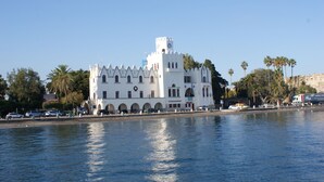 Un bâtiment blanc avec une tour horloge, situé au bord de l’eau, doté de fenêtres en arc de cercle et d’un balcon.