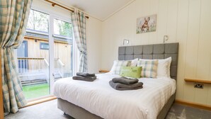 A bedroom with a large bed, a grey headboard, and a view of a wooden deck through a sliding glass door. - Robin Lodge (Skipton)