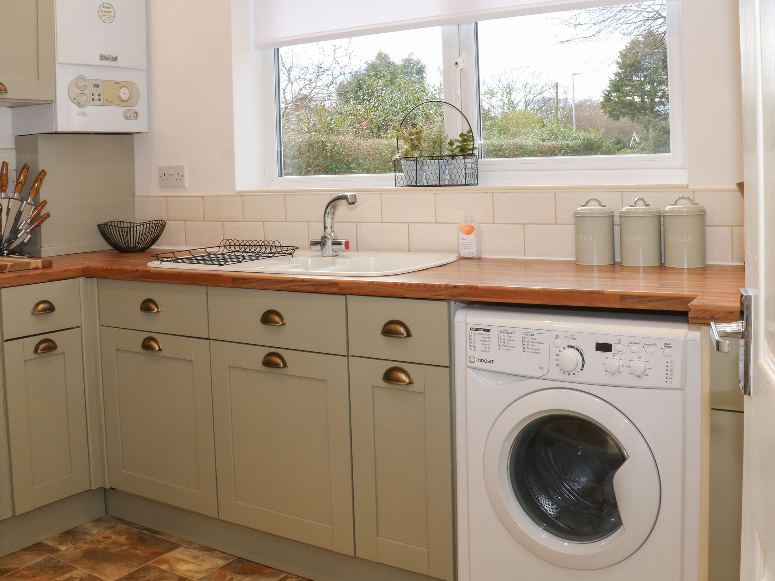 A kitchen with a built-in washing machine, a window with a view of greenery, and a sink with a soap dispenser.