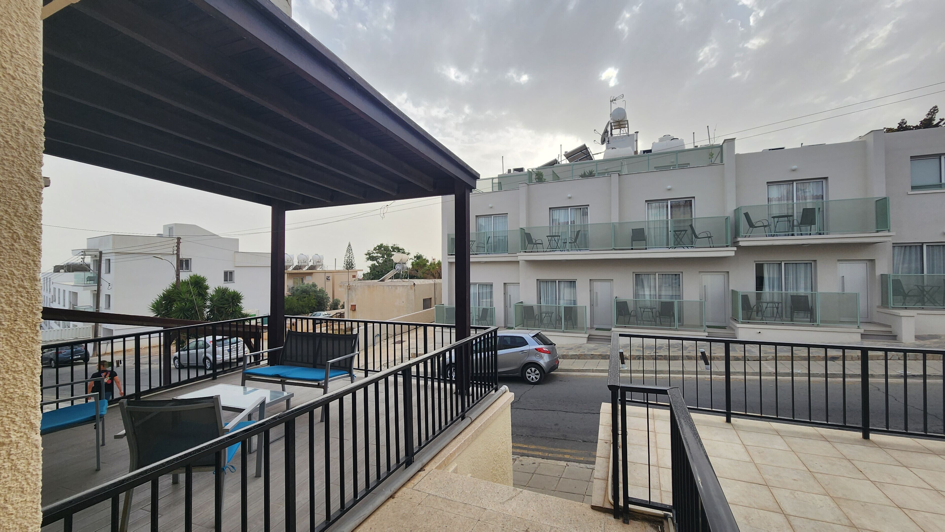 A balcony with outdoor seating, a car parked on the street, and a multi-story building in the background.
