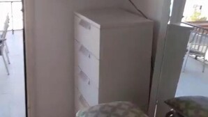 Quarto com televisão na parede, uma cadeira de madeira com almofada e um pequeno armário.