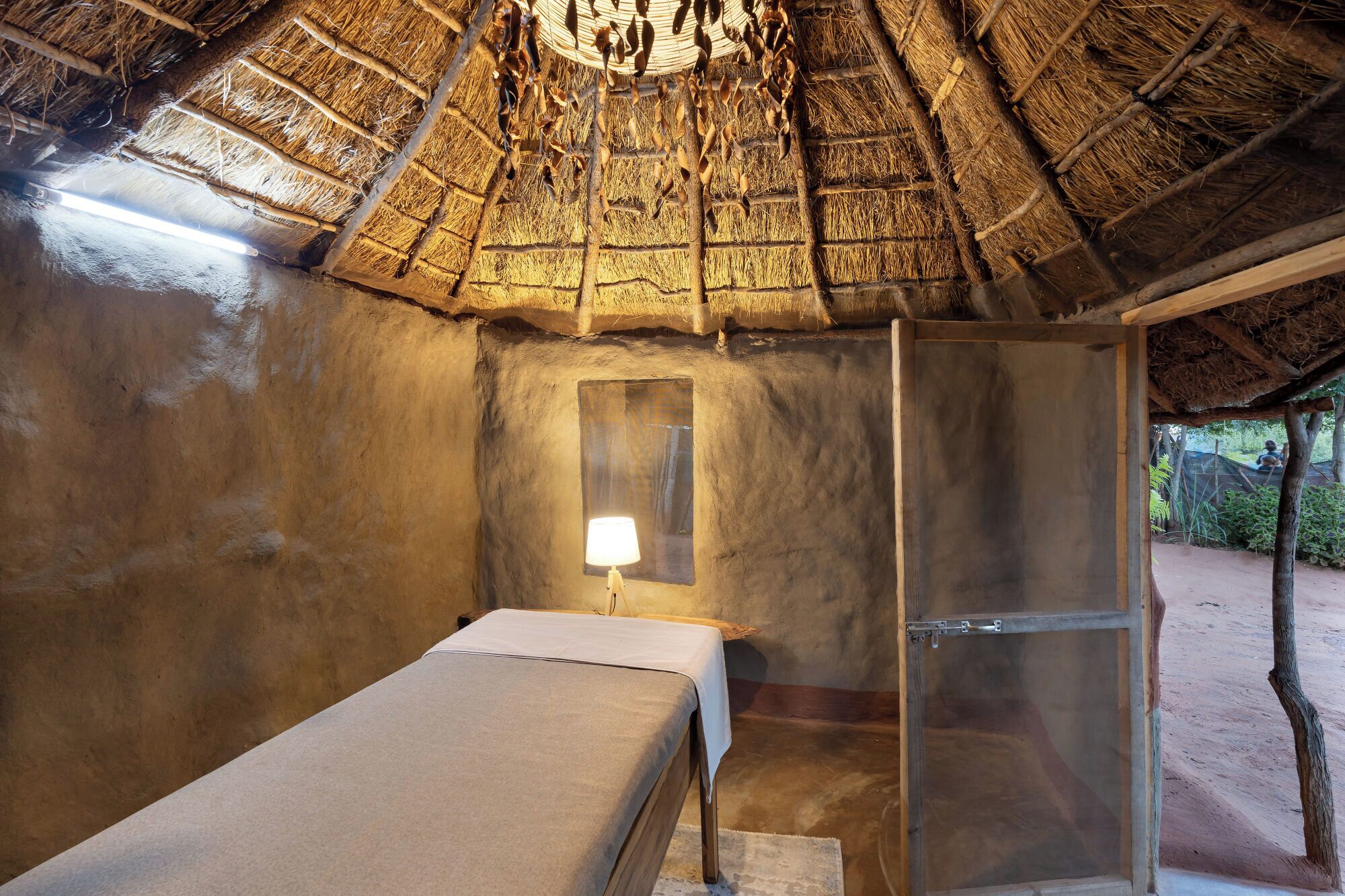 A rustic room with a thatched roof, a bed, a small window, and a door leading outside.