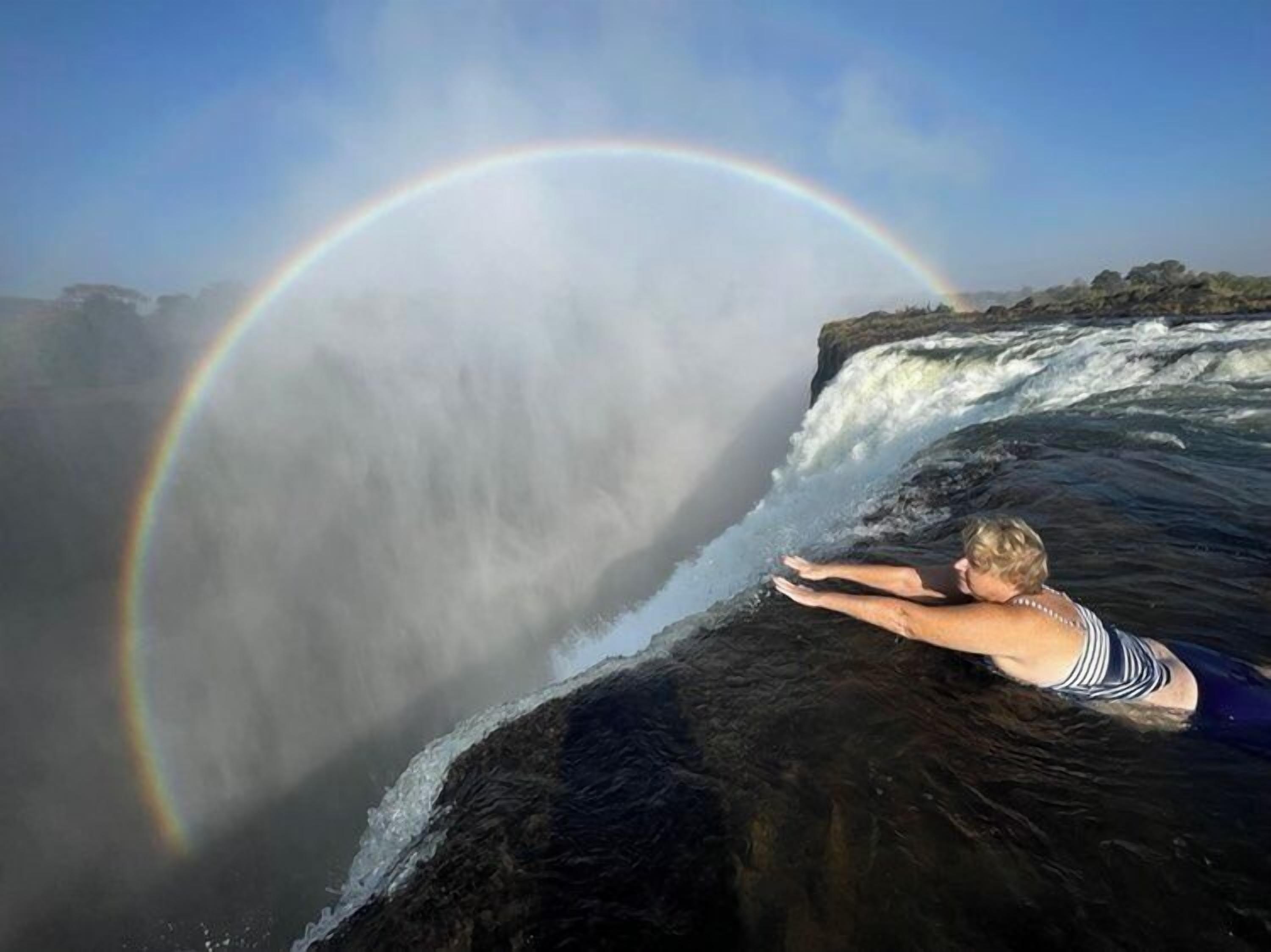 Une personne se détend sur un rocher près d’une cascade, avec un arc-en-ciel en arrière-plan.