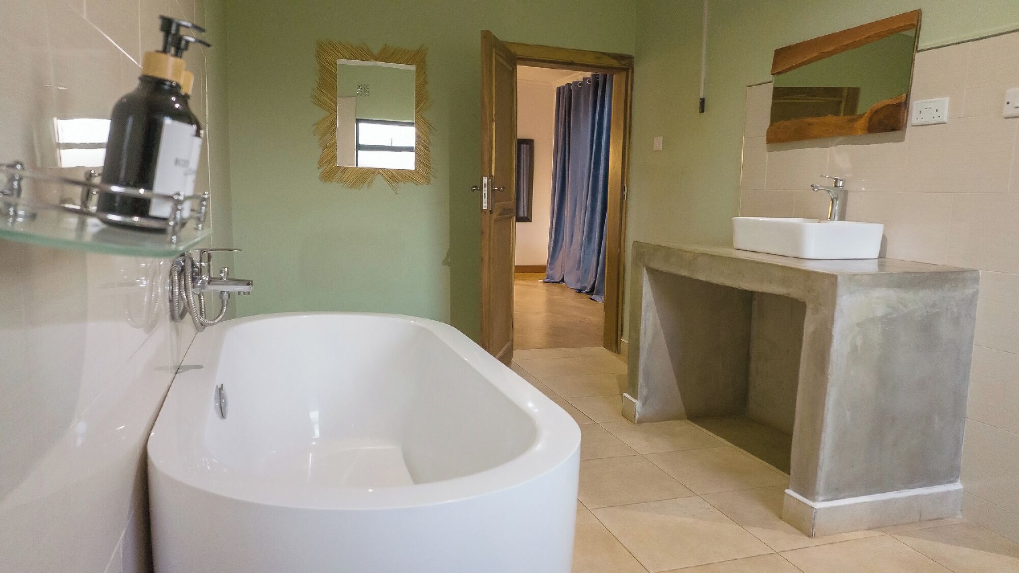 Un baño con una bañera independiente, un estante de vidrio con artículos de aseo, una puerta de madera que conduce a otra habitación y un tocador con espejo.