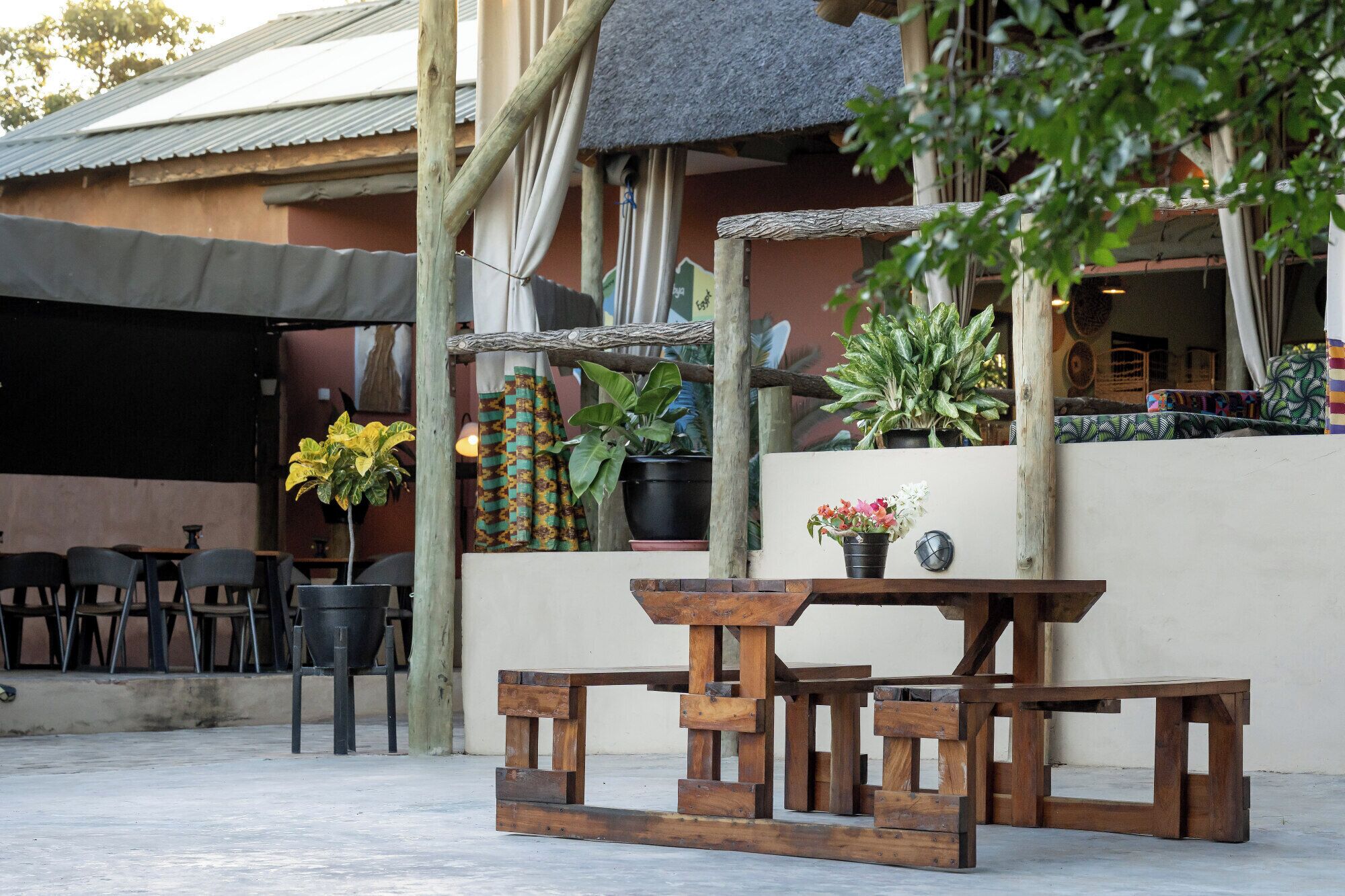 An outdoor seating area with wooden tables and chairs, surrounded by potted plants and a thatched roof structure.
