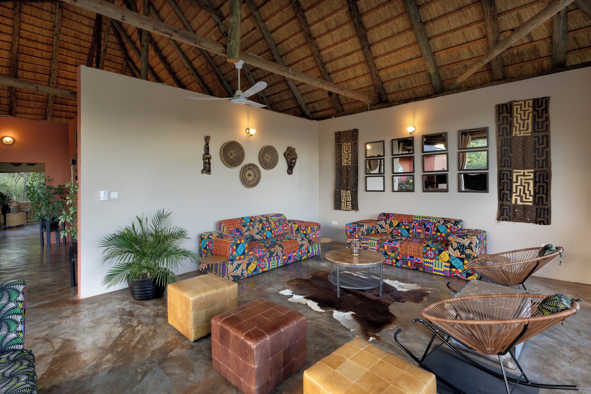 A spacious room with a high ceiling, a ceiling fan, and a thatched roof. There are two colorful sofas, a round coffee table, and a wicker chair. The walls are adorned with various decorative items and mirrors.