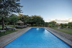 A swimming pool with lounge chairs and a thatched-roof structure, surrounded by greenery. - The Nkhosi Livingstone Lodge and Spa (Livingstone)