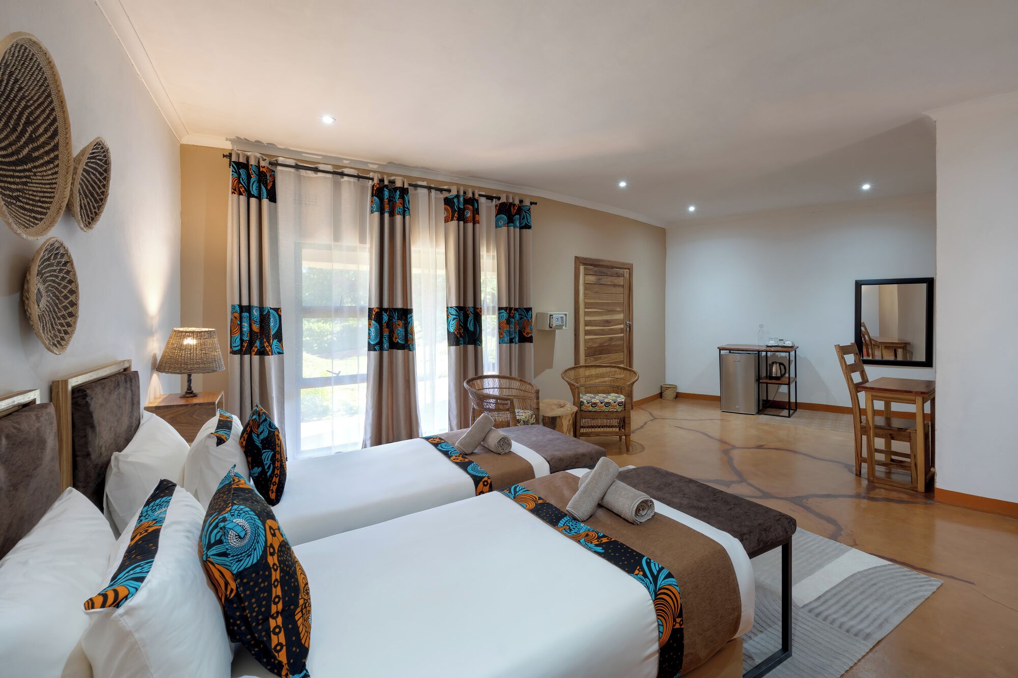 A hotel room with two beds, a desk, a chair, a mirror, and a small kitchenette.